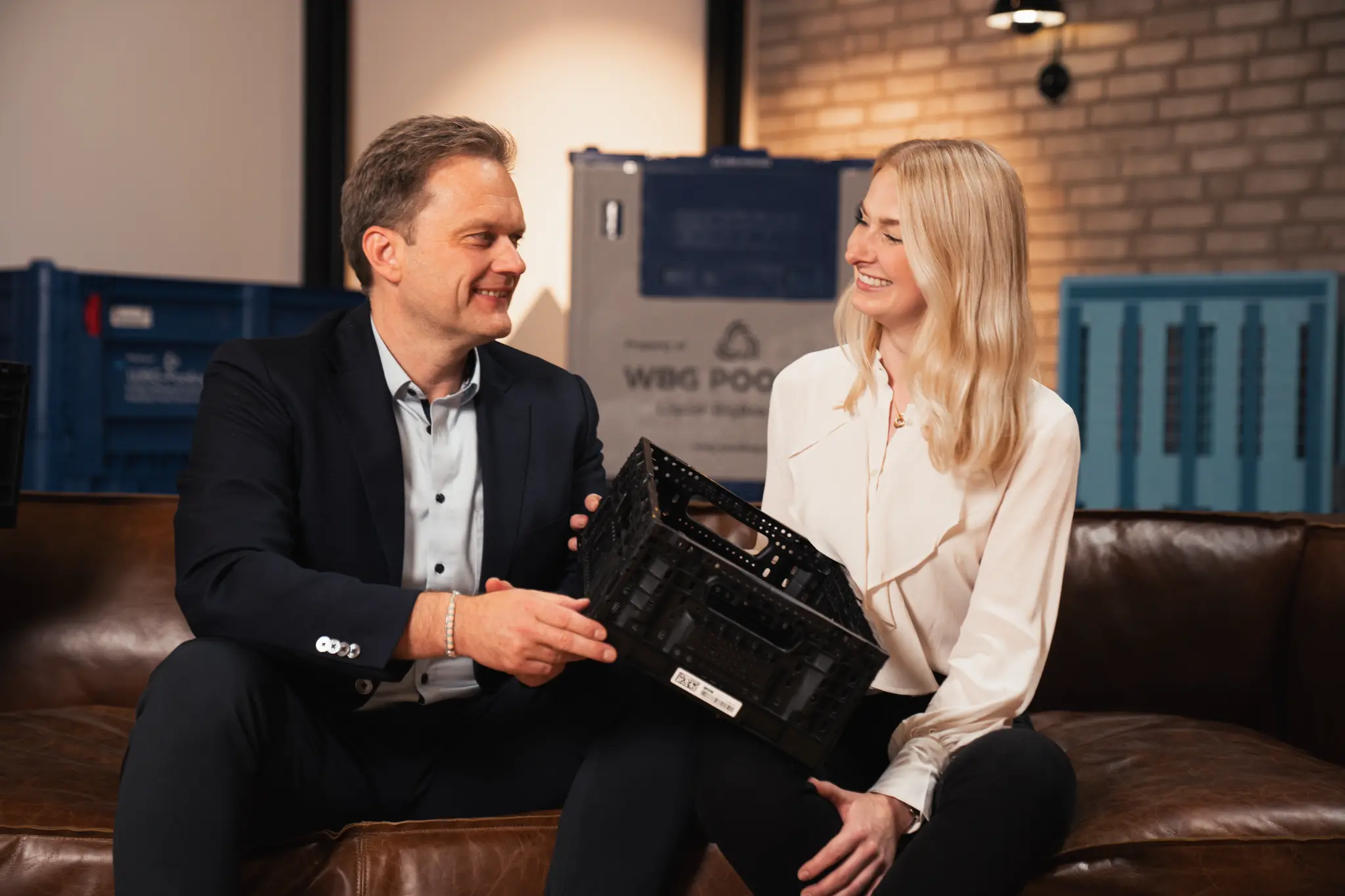 Zwei Mitarbeitende der Runden Group sitzen mit einer Mehrwegkiste auf einem Ledersofa.