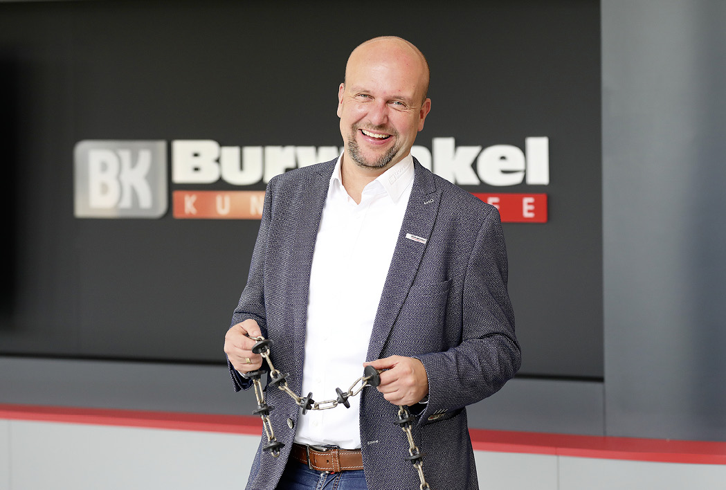 Martin Burwinkel steht vor dem Logo von Burwinkel Kunststoffwerk und hält eine Kunststoffkette in den Händen.