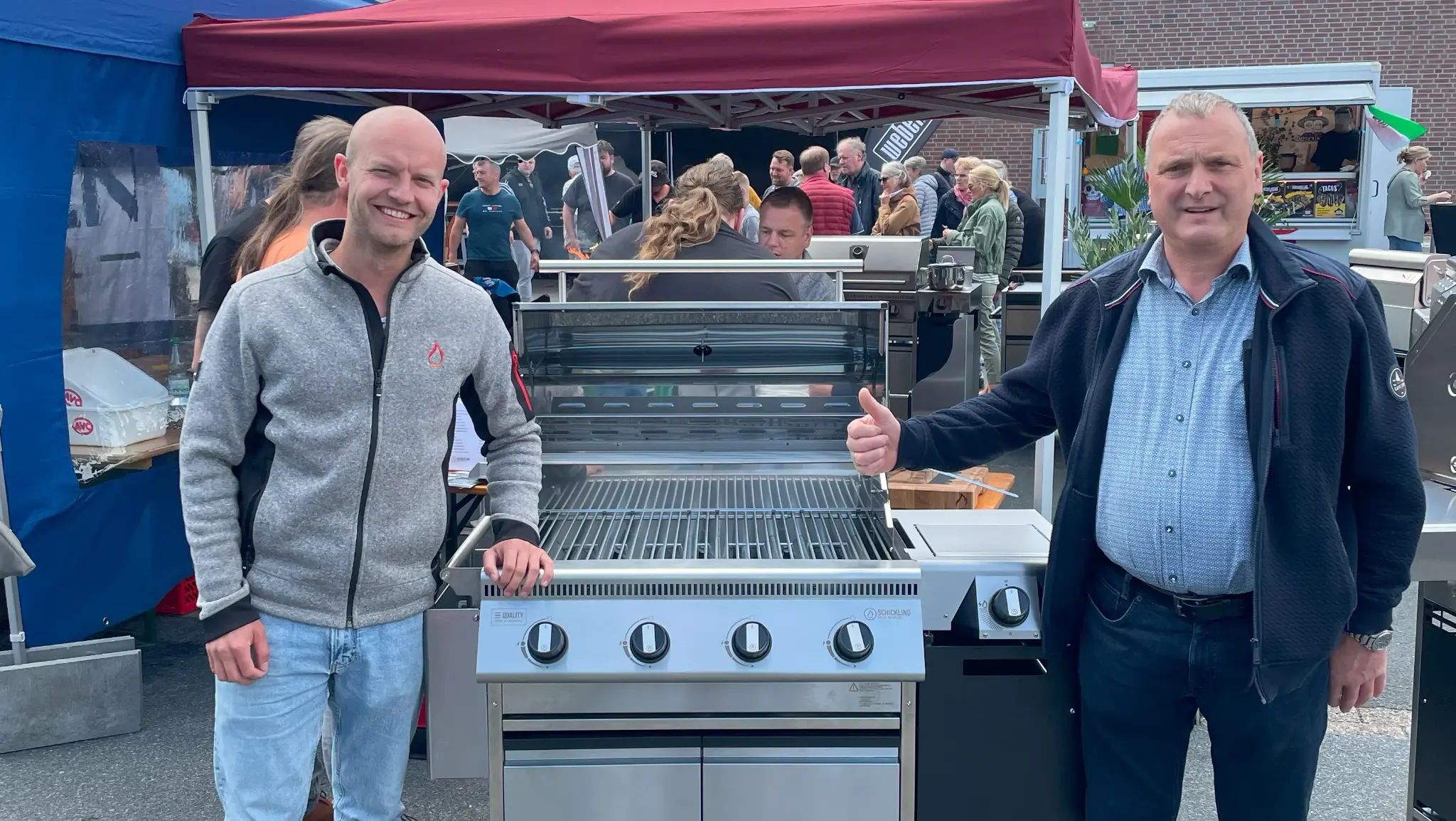 Zwei Personen stehen mit einem Schickling-Grill auf einer Outdoor-Veranstaltung vor einem Publikum.